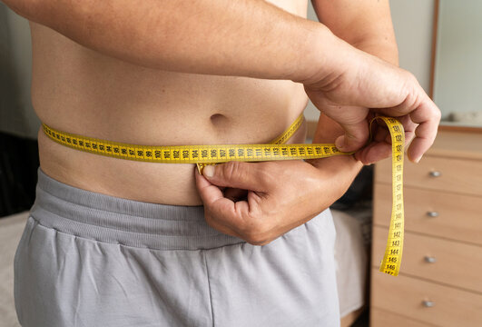Man measures his waist with tape measure at home, tracking his weight loss progress. Real person, self-acceptance, natural lifestyle, weight control