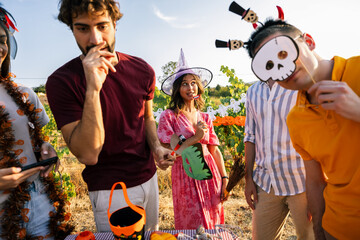Obraz premium Young people in Halloween costumes holding skull and monster photo props at a party table.