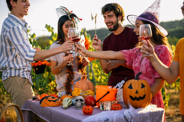 Obraz premium Group of joyful young friends wearing Halloween costumes and toasting with red wine outdoors at a vineyard party.