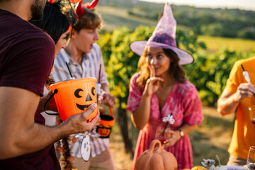 Obraz premium Woman in a witch hat making a shushing gesture while friends gather with a Halloween bucket in a vineyard.