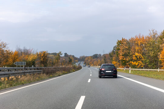 Back scenic view black family car driving highway road through bright autumnal golden red orange nature foliage forest trees. Scenery fall season driveway vehicle. Drive auto safety