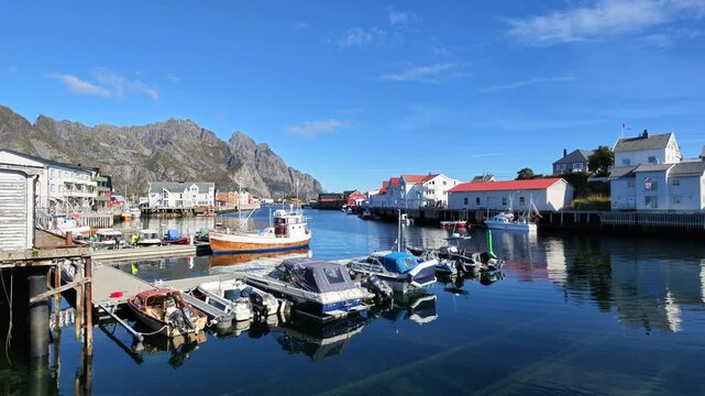 Sunny harbor scene in Henningsvaer on the Lofoten Islands with numerous boats, traditional houses, calm blue water and impressive rocky mountains under clear sky.