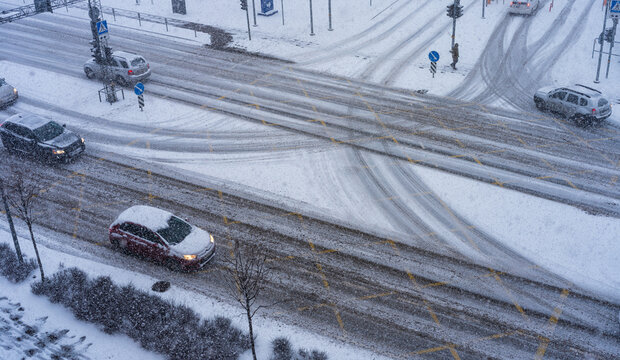 View of a snow-covered intersection in Vilnius with cars driving through winter weather, featuring tire tracks on slushy roads and traffic lights in Vilnius, Vilnius County, Lithuania.