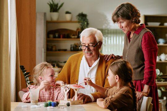 Senior Caucasian man with mature woman watching two little girls painting wooden craft at home. Scene highlighting family connection for intergenerational lifestyle advertising