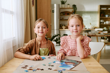 Obraz premium Portrait of two Caucasian female children sitting at table, assembling jigsaw puzzle at home. Image suitable for family leisure, early childhood development, cognitive gaming campaigns