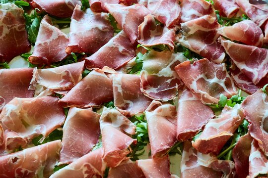 Slices of Italian capocollo ham on a bed of fresh green arugula. Gourmet bresaola or prosciutto cold cuts, traditional Mediterranean antipasto meat platter, catering food background.