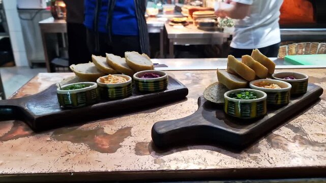 Sliced artisan baguette bread rests on dark wooden boards beside ramekins of savory dips and green scallions. Commercial kitchen counters and busy chefs work in the background.