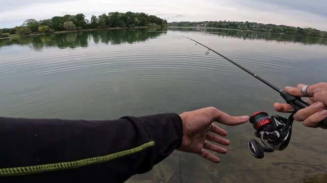 POV fisherman casting fishing rod with spinning reel on calm lake water. Relaxing outdoor freshwater angling and nature recreation.