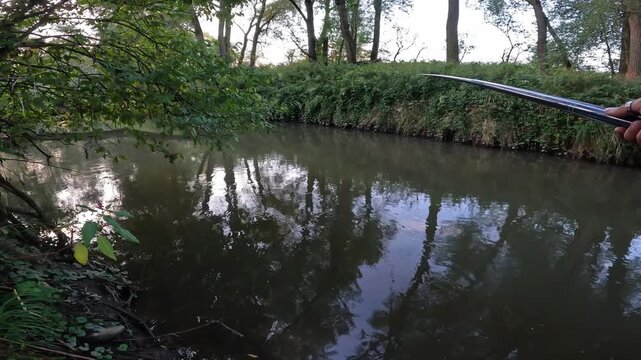 POV of an angler holding a long fishing pole rod over a calm forest river. First person view of summer freshwater angling.
