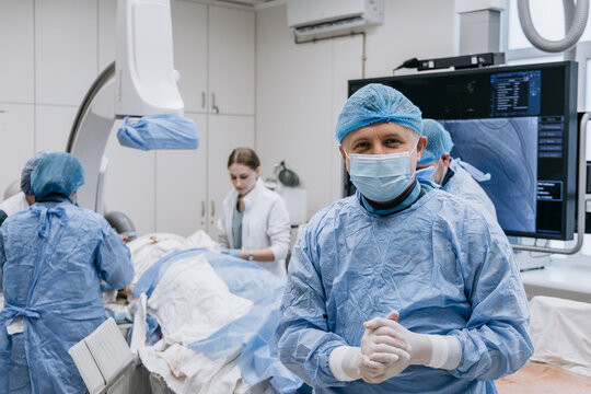 Cardiologist looking at the camera with a patient and medical team working in the background.