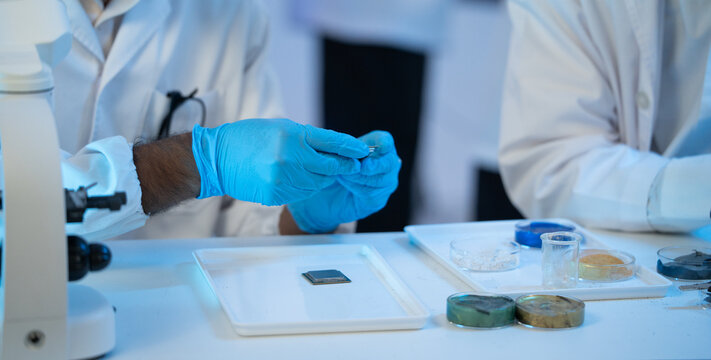 A team of researchers examines a computer chip and chemical samples at a table inside a laboratory, focusing on precise analysis and technical problem solving during an experiment.