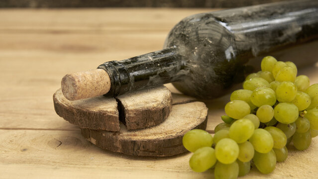 A fragment of a dusty dark-glass wine bottle lying on its side, with a bunch of light-colored grapes nearby against a wooden background.