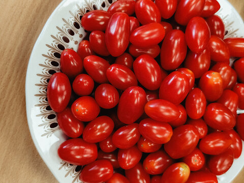 Ripe red grape tomatoes piled on a decorative ceramic plate with varied oval shapes and glossy skins, photographed from above on a wooden tabletop surface
