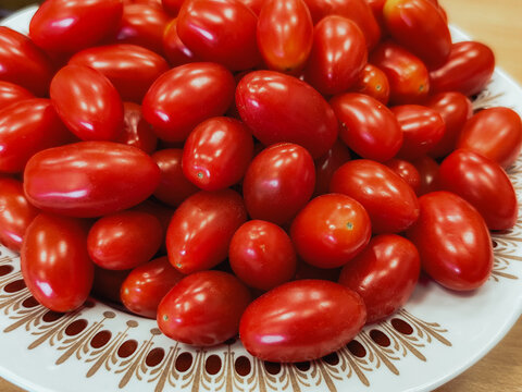 Fresh red oval tomatoes piled on a decorative white ceramic plate, showing glossy skins and natural variations in shape and size, food still life