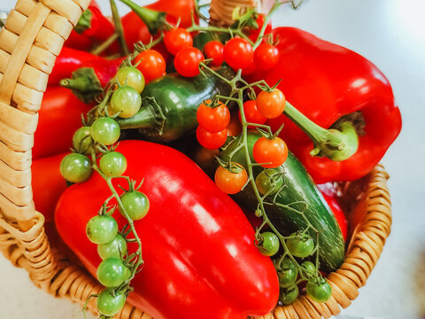 Fresh red peppers, cucumbers, and green and orange tomatoes arranged in a woven basket with vibrant colors and natural textures, overhead produce still life