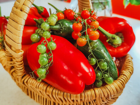 Fresh red peppers, cucumbers, tomatoes, and green fruits arranged in a woven basket, colorful close-up of assorted garden produce on a light surface