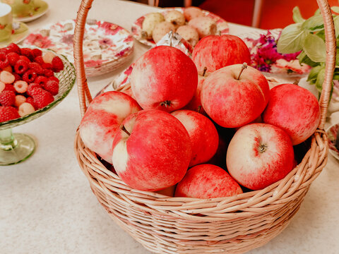 Basket filled with red apples beside raspberries and floral dishes on a decorated dining table, colorful seasonal still life with fresh fruit and woven centerpiece