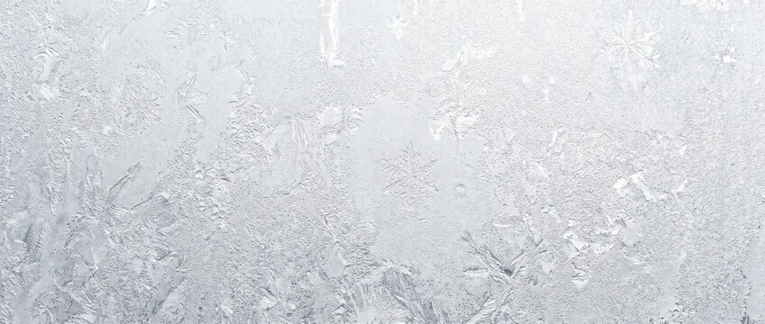 Frosted glass texture with icy snowflake patterns. Close-up of frozen winter window surface. Bright white cold weather holiday background