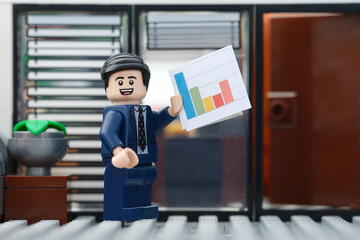 Fototapeta premium Tambov, Russian Federation - September 23, 2026 A happy Lego businessman minifigure standing in an office setting while holding up a colorful bar chart document.