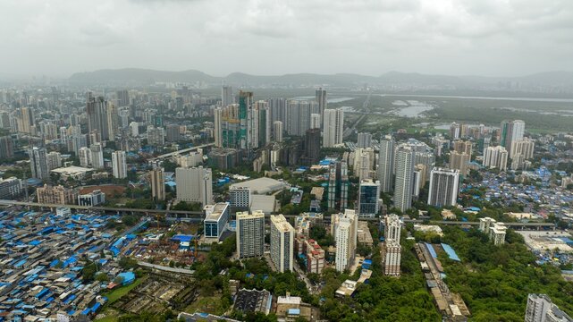 Aerial view of Dharavi Slums with its dense blue-roofed structures contrasting with the modern high-rise skyline and lush green patches in the background in Mumbai, Maharashtra, India.