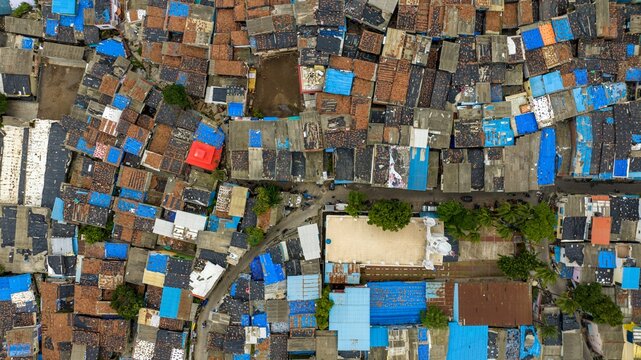 Aerial view of the dense Dharavi Slums featuring a patchwork of corrugated metal and blue plastic roofs, narrow alleyways, and a white-roofed building in Mumbai, Maharashtra, India.