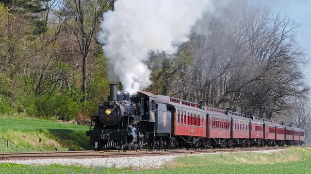 Ronks, Pennsylvania, United States - 08 April 2026: View of a historic steam train pulling red passenger cars through a lush green landscape with trees.