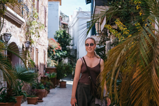 Young woman exploring sunny Mediterranean village streets with white houses, flowers and tropical plants, capturing solo travel, vacation lifestyle and relaxed summer wanderlust.