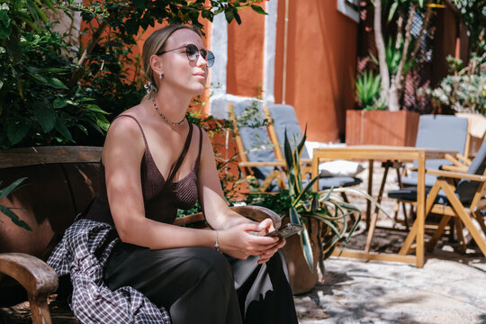 Young woman using smartphone in cozy Mediterranean cafe and garden courtyard, showing solo travel, vacation lifestyle, leisure, mobile connection and relaxed summer tourism.