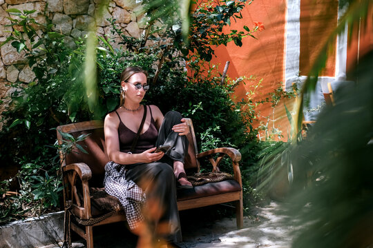 Young woman using smartphone in cozy Mediterranean cafe and garden courtyard, showing solo travel, vacation lifestyle, leisure, mobile connection and relaxed summer tourism.