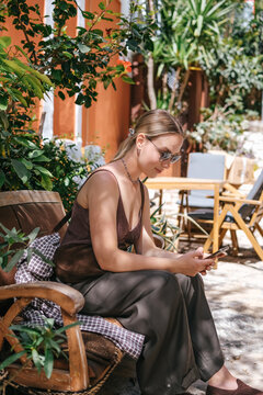 Young woman using smartphone in cozy Mediterranean cafe and garden courtyard, showing solo travel, vacation lifestyle, leisure, mobile connection and relaxed summer tourism.