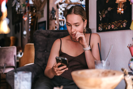 Young woman using smartphone in cozy Mediterranean cafe and garden courtyard, showing solo travel, vacation lifestyle, leisure, mobile connection and relaxed summer tourism.
