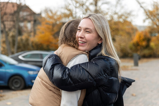 Smiling woman hugs friend outdoors during candid autumn meeting, expressing joy, affection and emotional connection. Urban background and fall colors suggest reunion, greeting or emotional goodbye