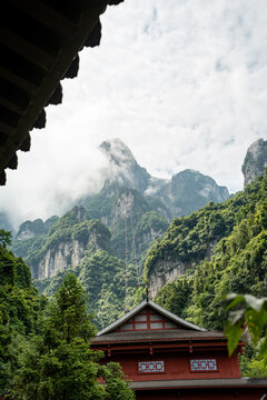 A modern cable car steeply ascends into mist-shrouded karst peaks, while a traditional Chinese building in the foreground frames the majestic scene.