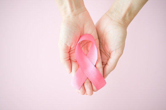 Woman holding pink ribbon in hands for breast cancer awareness on pale pink background.