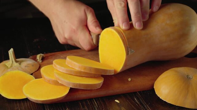 Hands slicing a butternut squash into round pieces with a kitchen knife on a wooden cutting board, surrounded by pumpkin sections and seeds in a rustic kitchen setting