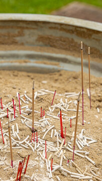 Incense sticks of various lengths inserted into sandy soil within a round metal container, surrounded by a green outdoor setting