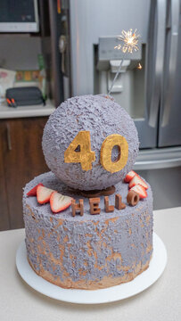 Birthday cake with purple frosting, topped with a spherical decoration, gold number 40, and strawberries, set on a kitchen countertop with appliances in the background