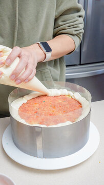 Person in green sweatshirt decorates cake with piping bag, filling a round cake mold on a kitchen countertop with stainless steel appliances in the background