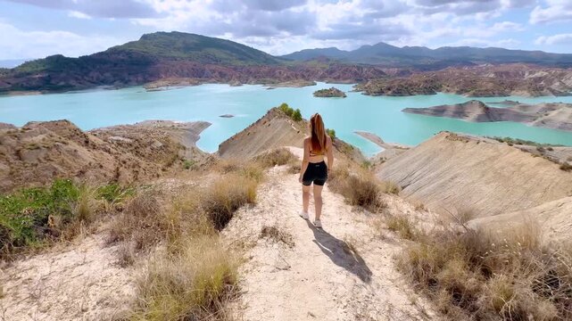 Cinematic Rear View of Woman in Blue Dress Walking Across Desert Hills Toward Turquoise Lake at Barranco de Gebas Murcia Spain