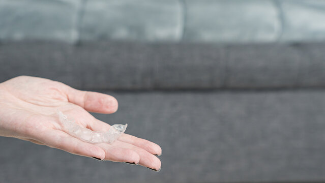 Hand holding a transparent dental retainer above a gray sofa with a textured fabric surface in a modern living room setting