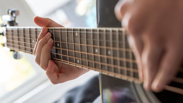 Close-up of hands playing acoustic guitar, focusing on the fretboard and strings, with a bright window in the background illuminating the scene