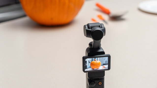 Camera capturing a close-up of a carved pumpkin on a kitchen countertop with orange carving tools and a spoon visible in the background