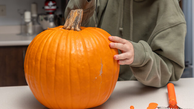 Child carving a pumpkin for Halloween with a knife, wearing a green hoodie, orange pumpkin on a kitchen countertop with carving tools nearby