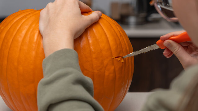 Person carving an orange pumpkin for Halloween using a small serrated knife on a kitchen countertop with blurred background elements