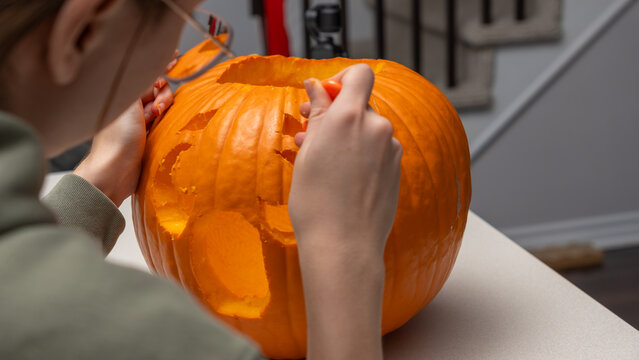 Person carving a Halloween pumpkin with intricate designs, seated at a table in a well-lit indoor space with stairs visible in the background