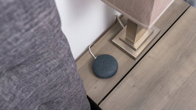Smart home wireless charger on wooden nightstand beside a modern lamp and gray upholstered headboard in a contemporary bedroom setting