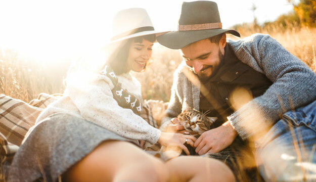 Young couple spends time together sitting on blanket in field, petting a cat. Couple enjoys relaxing outdoors. Concept of love, enjoyment, and day off.