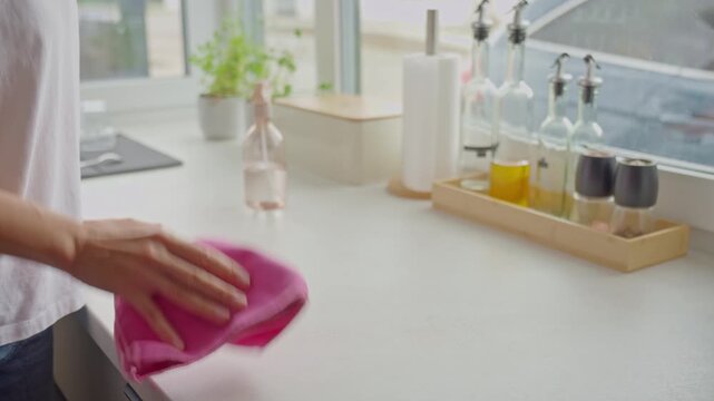 Woman sprays cleaner onto kitchen countertop and wipes surface with pink cloth. Household cleaning and kitchen hygiene