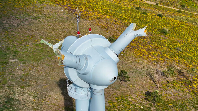 Aerial view of a damaged wind turbine with broken blades and structural damage set against a vibrant yellow flowering hillside in Lisbon, Lisbon, Portugal.