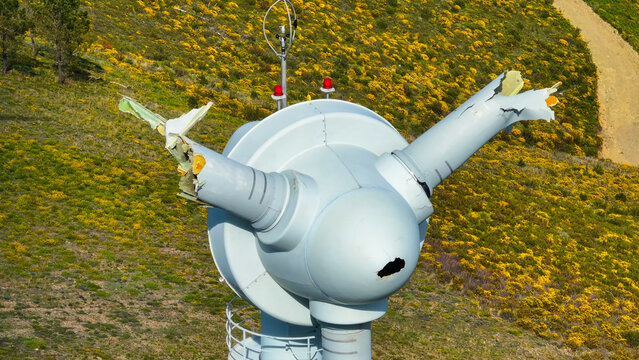 Aerial view of a damaged wind turbine hub with broken blades and structural failure set against a vibrant yellow flowering hillside in Lisbon, Lisbon, Portugal.